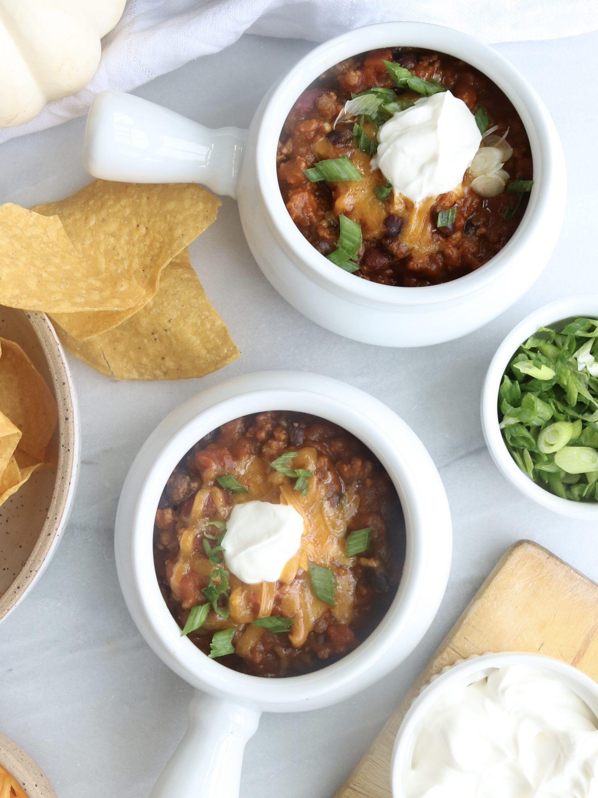 two bowls of pumpkin chili, corn chips, and scallions on a white surface